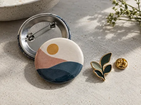 A round button badge with visible safety-pin back next to a small enamel pin with its clutch backing, original abstract designs only.