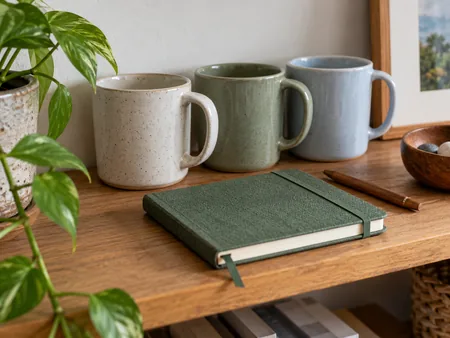 Three plain similar ceramic mugs together on a shelf, with a different small useful notebook in foreground as an alternative gift idea.