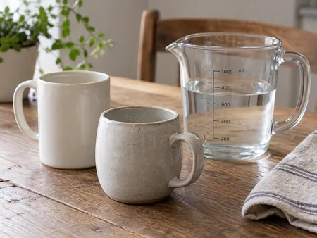 Two differently shaped plain ceramic mugs beside a transparent graduated measuring jug with water, three-quarter tabletop view.