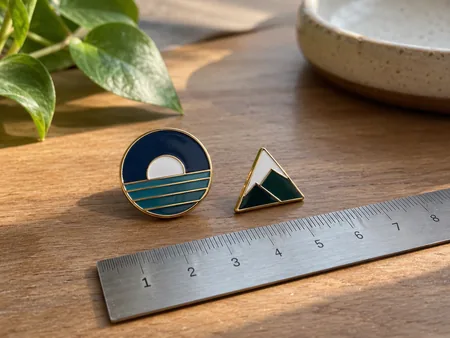 Two original simple geometric enamel pins of different sizes beside an ordinary ruler for scale, no recognised characters or letters.