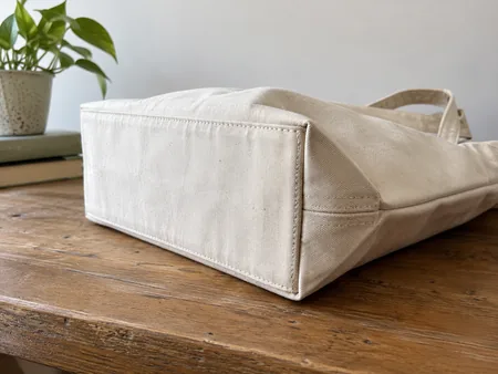 Underside and lower corner of a plain canvas tote shown on its side, reinforced rectangular base and stitched corner visible.